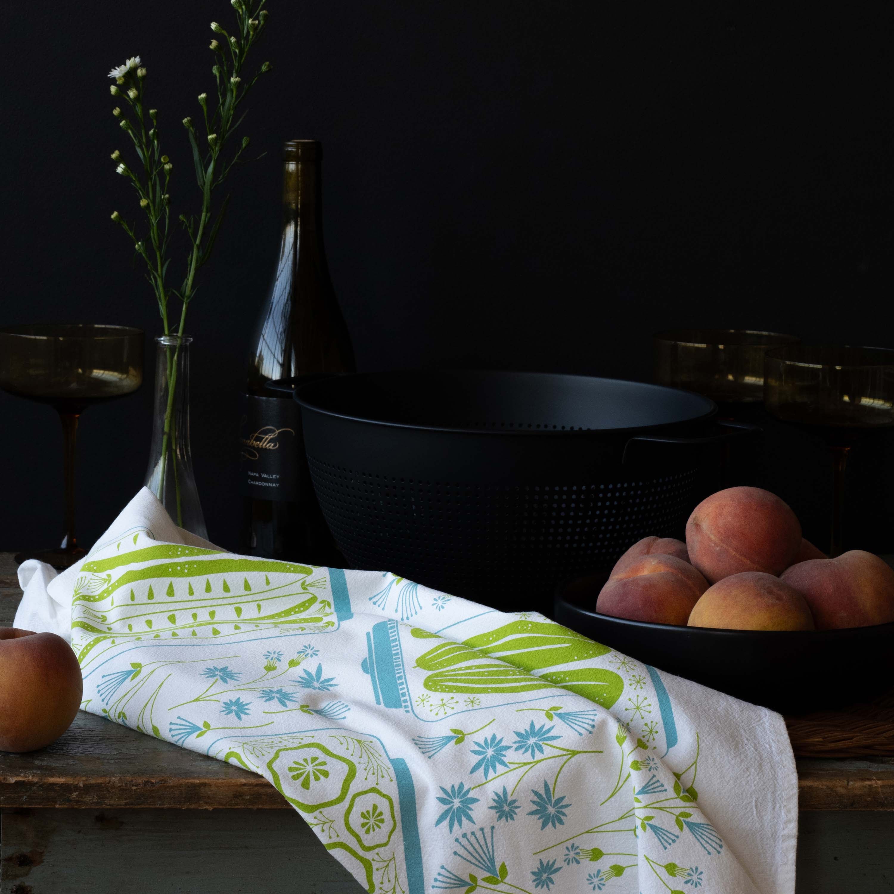 Pickles + Petals Tea Towel styled on a rustic table with peaches, a wine bottle, and dark cookware