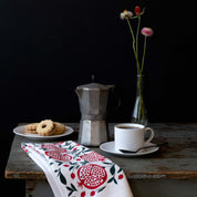 Pomegranates Tea Towel styled on a dark table with a moka pot, coffee, pastries, and a vase