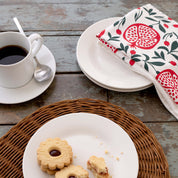 Pomegranates Tea Towel styled beside a coffee cup, plates, and cookies on a rustic tabletop