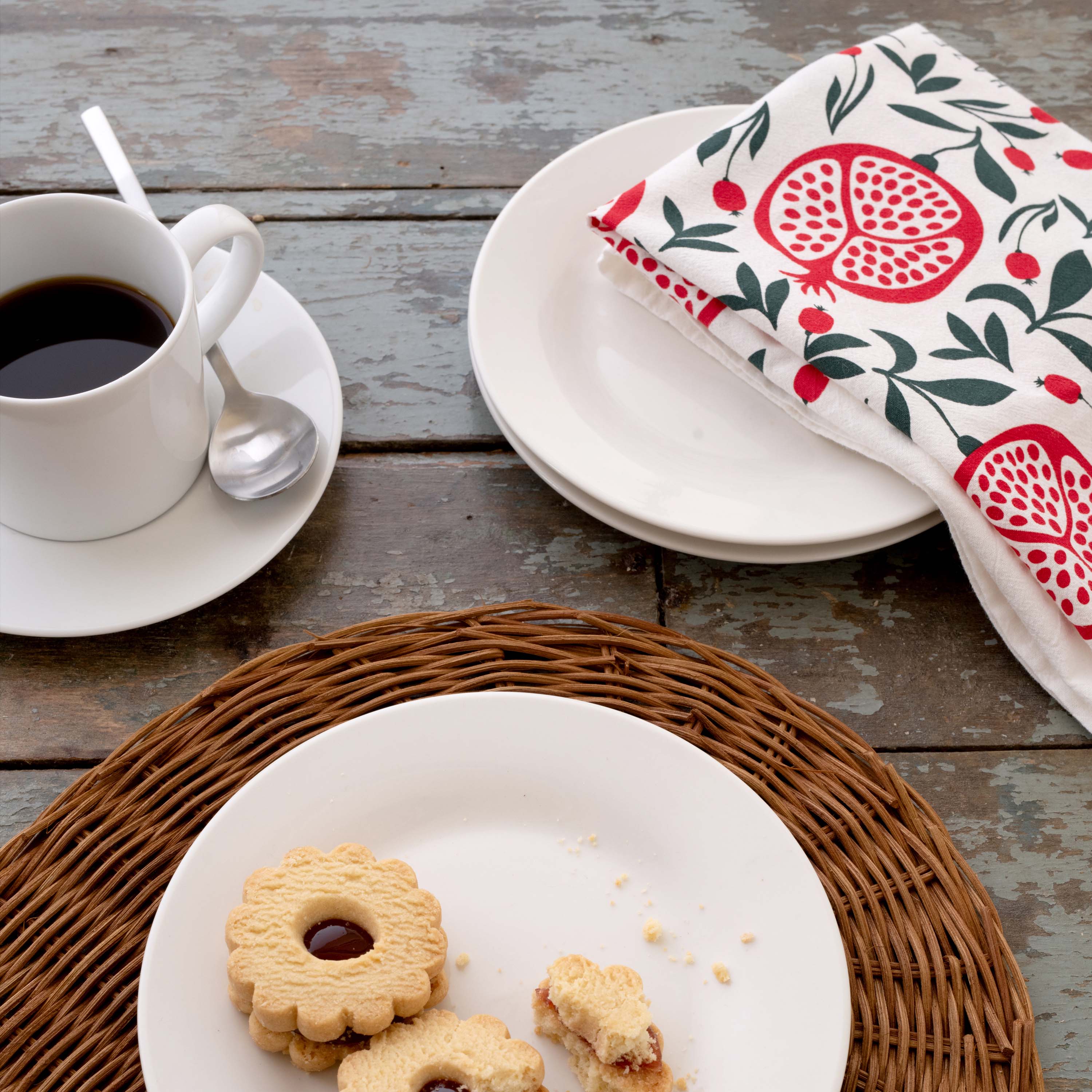 Pomegranates Tea Towel styled beside a coffee cup, plates, and cookies on a rustic tabletop