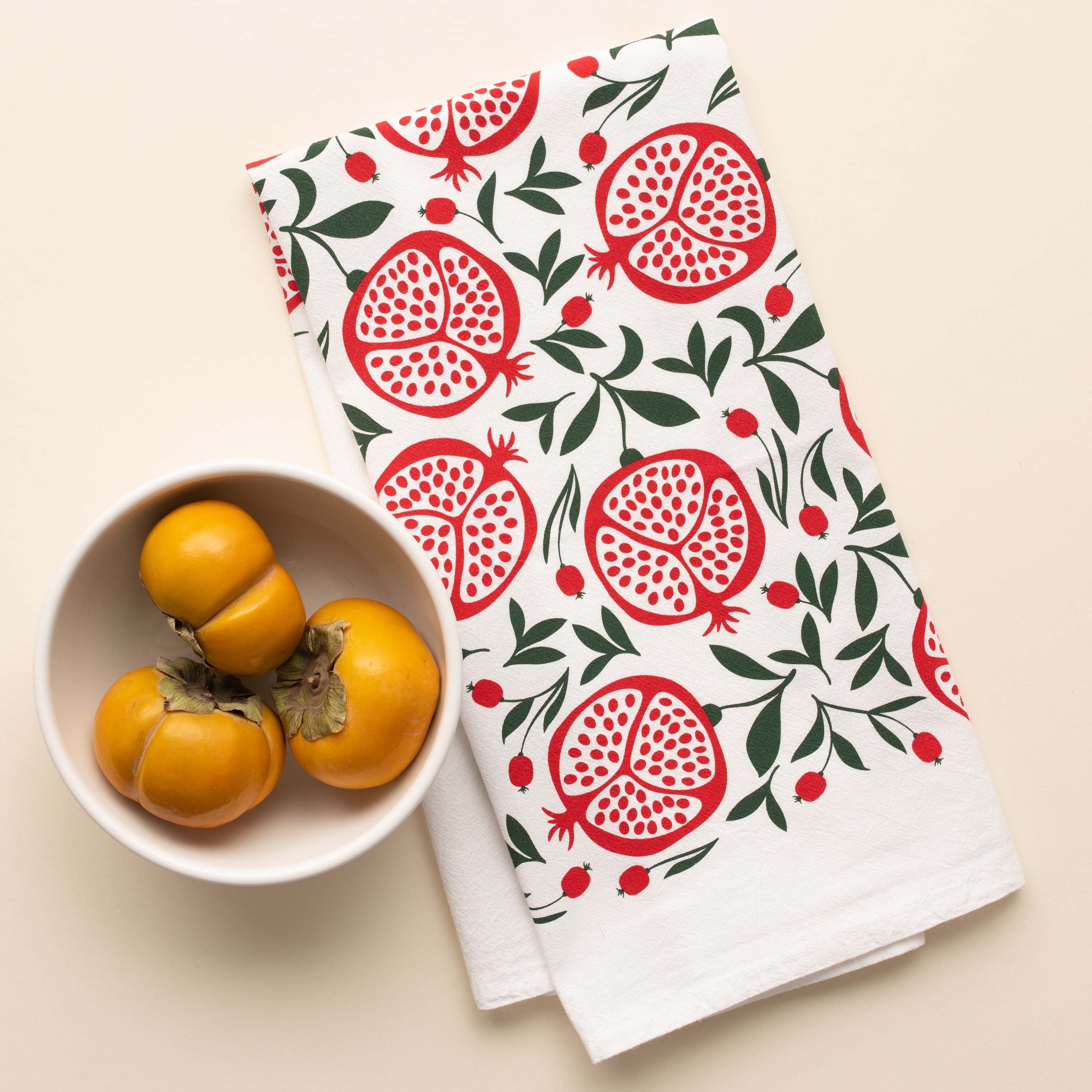 Pomegranates Tea Towel with hand-illustrated pomegranate design beside a bowl of yellow tomatoes