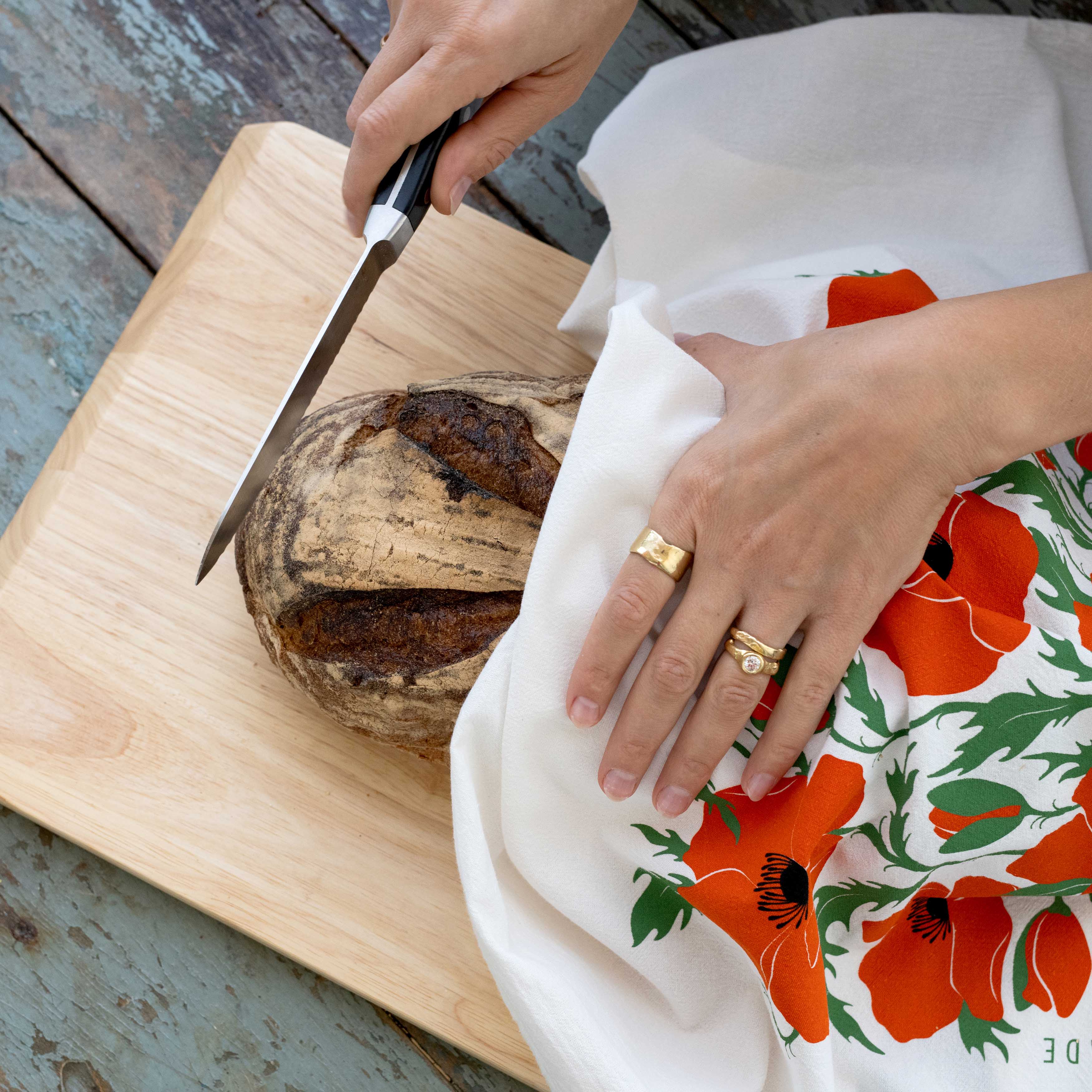 Hands slicing bread with a hand-illustrated poppy tea towel on a wooden board