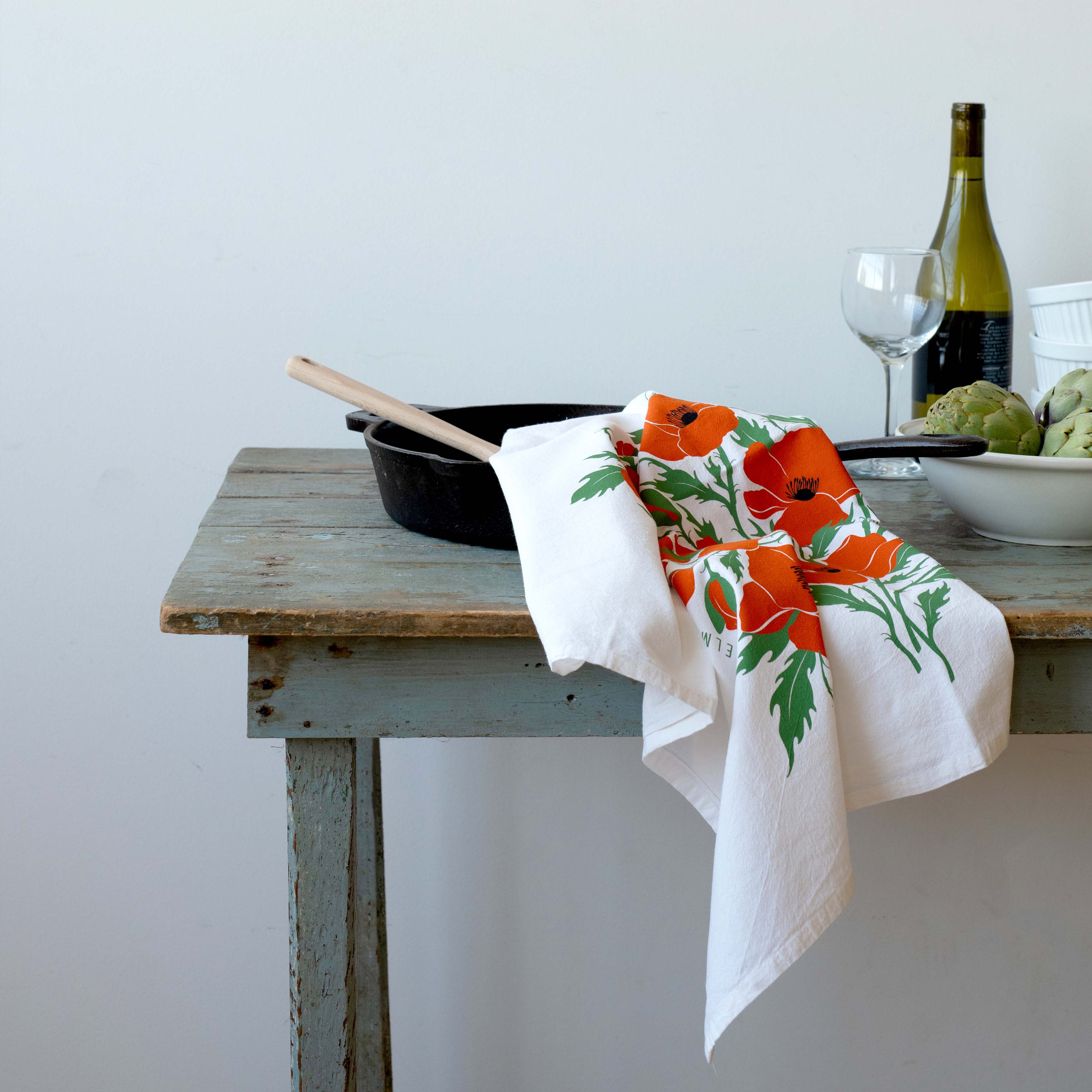 Poppies Tea Towel draped over a kitchen counter beside a skillet, wine bottle and artichokes