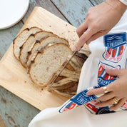 Sardines Tea Towel wrapped around sliced bread on a cutting board while someone slices the loaf