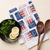 Sardines Tea Towel with hand-illustrated sardine tin design beside lemon slices, herbs, and wooden kitchen utensils
