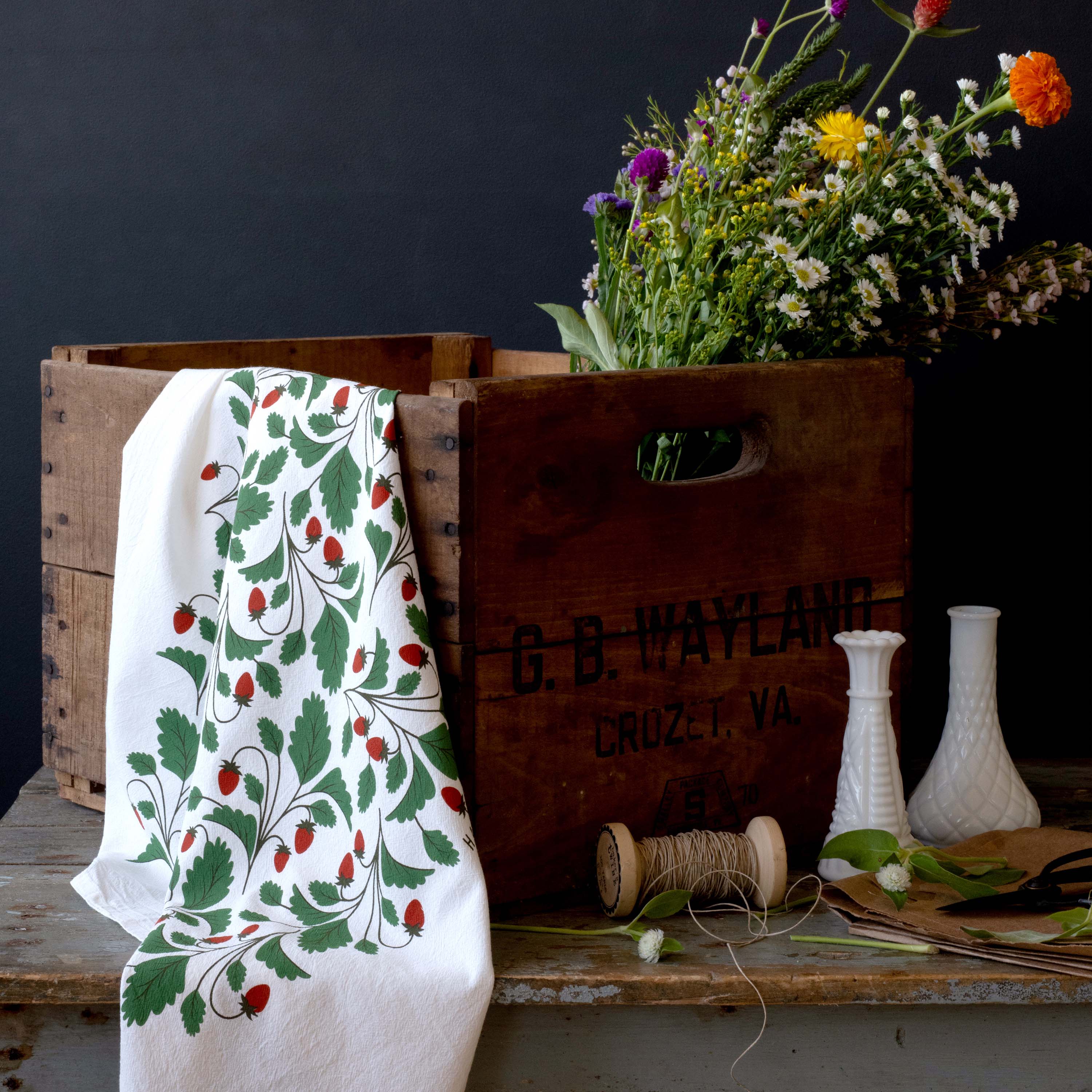Wild Strawberries Tea Towel draped over a wooden crate with wildflowers, and glass vases