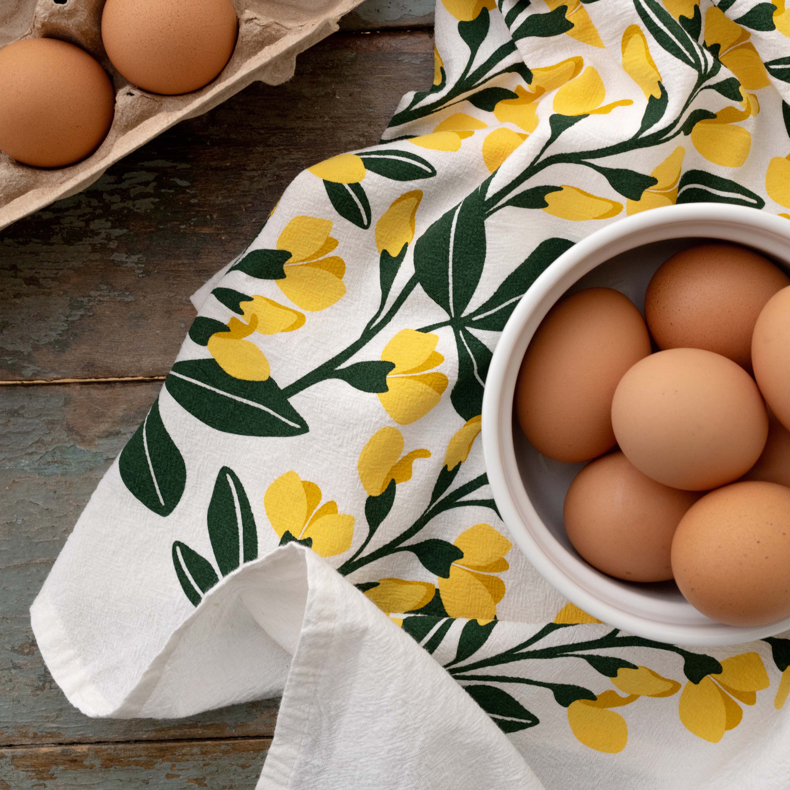 Sundrops Tea Towel draped beside a bowl of brown eggs on a wooden table
