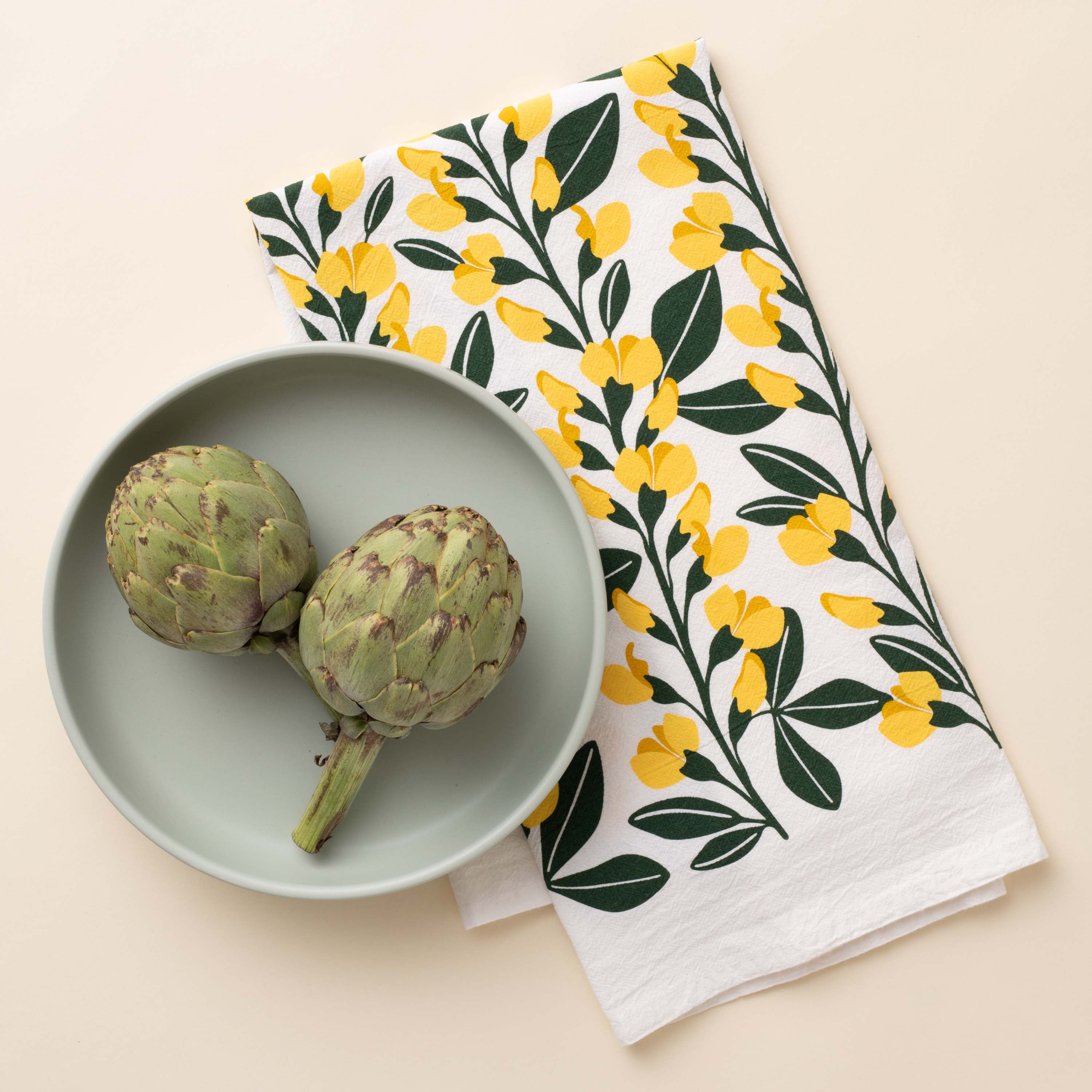Sundrops Tea Towel with hand-illustrated yellow floral design beside a plate with artichokes