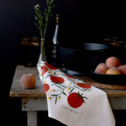 Tomatoes Tea Towel styled on a rustic table with peaches, a glass vase, and dark cookware
