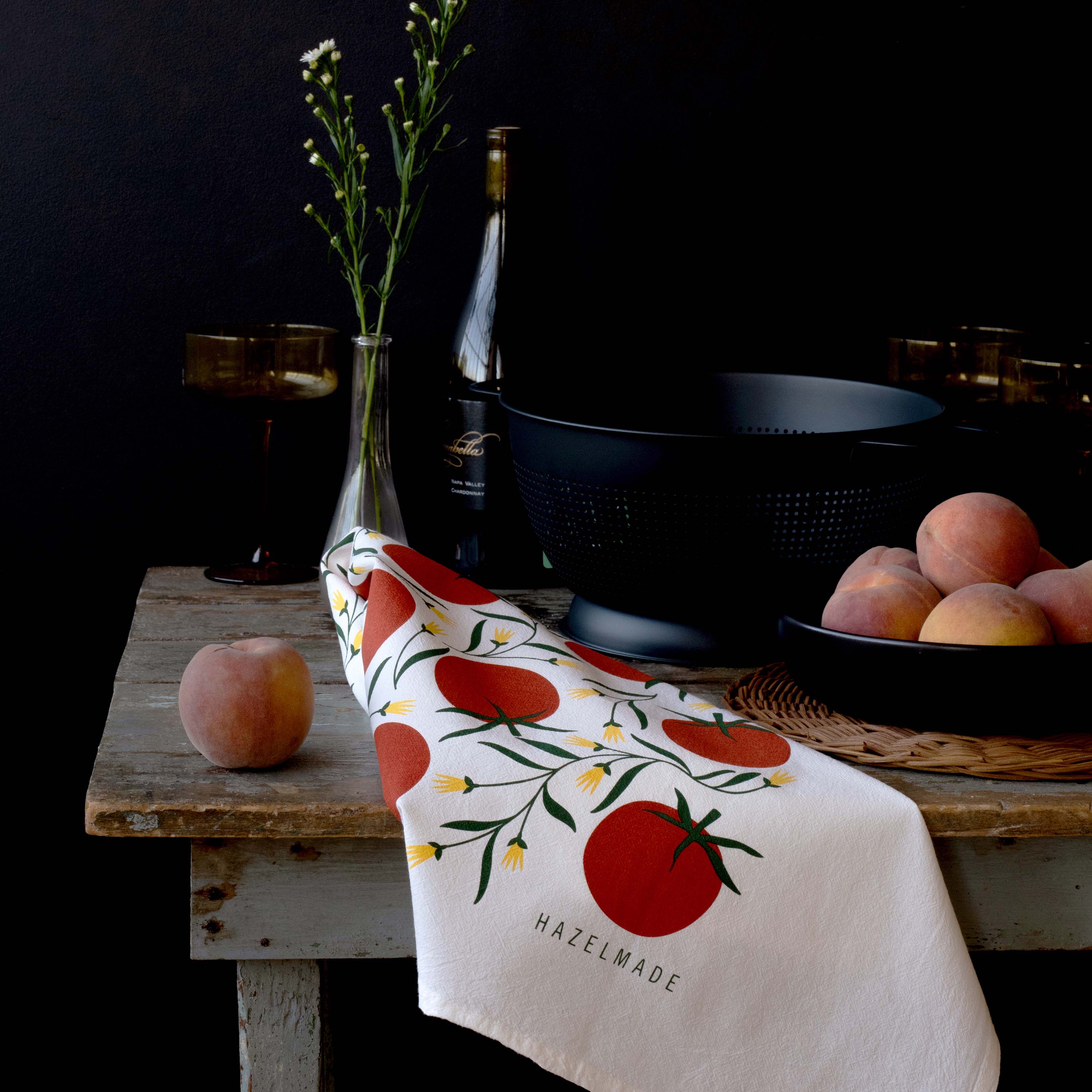 Tomatoes Tea Towel styled on a rustic table with peaches, a glass vase, and dark cookware