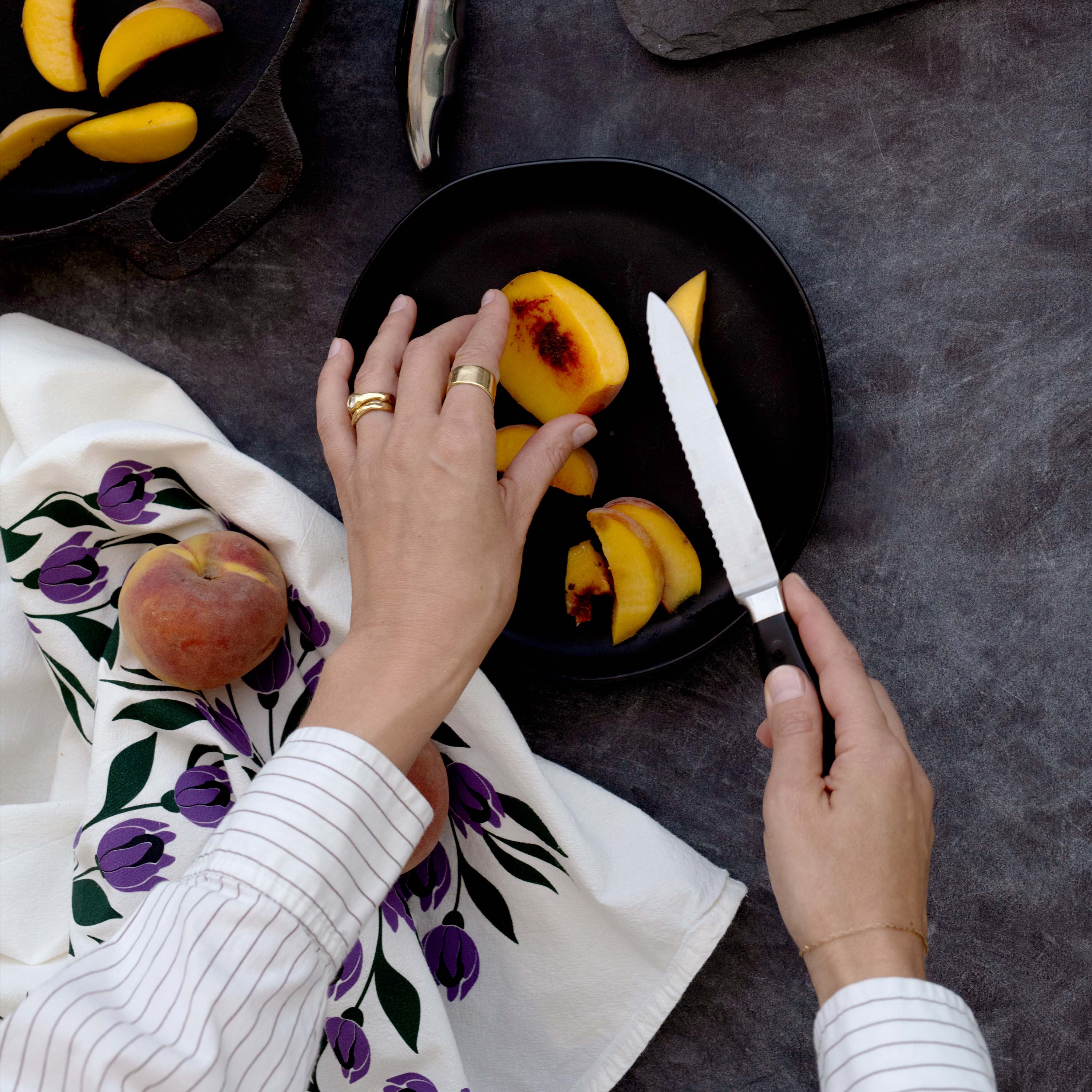 Tulips Tea Towel sitting on a dark tabletop while someone slices peaches on a black plate