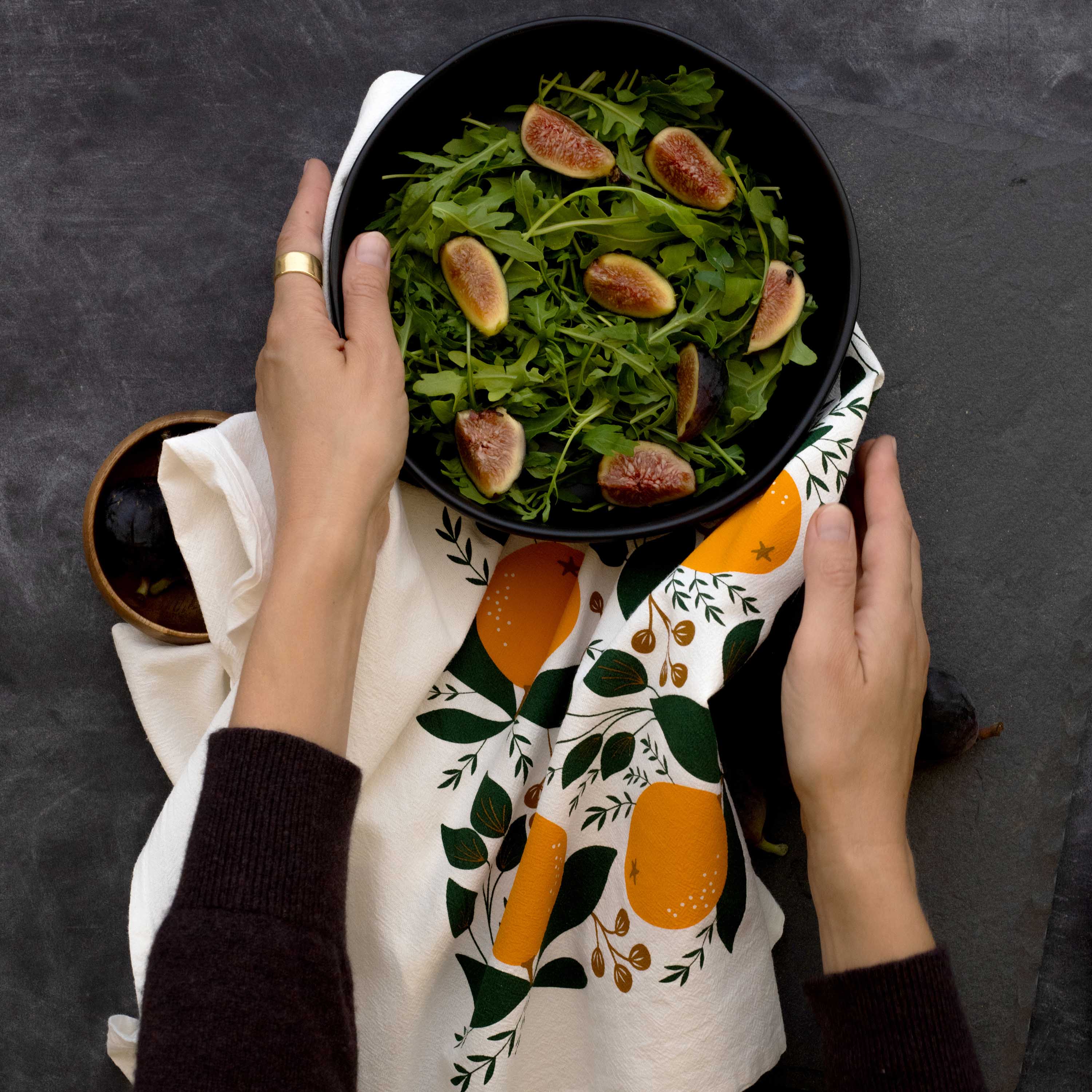 Hands holding a bowl of fig salad over a hand-illustrated citrus tea towel