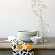 Winter Citrus Tea Towel styled beside stacked bowls and a glass vase on a wooden table