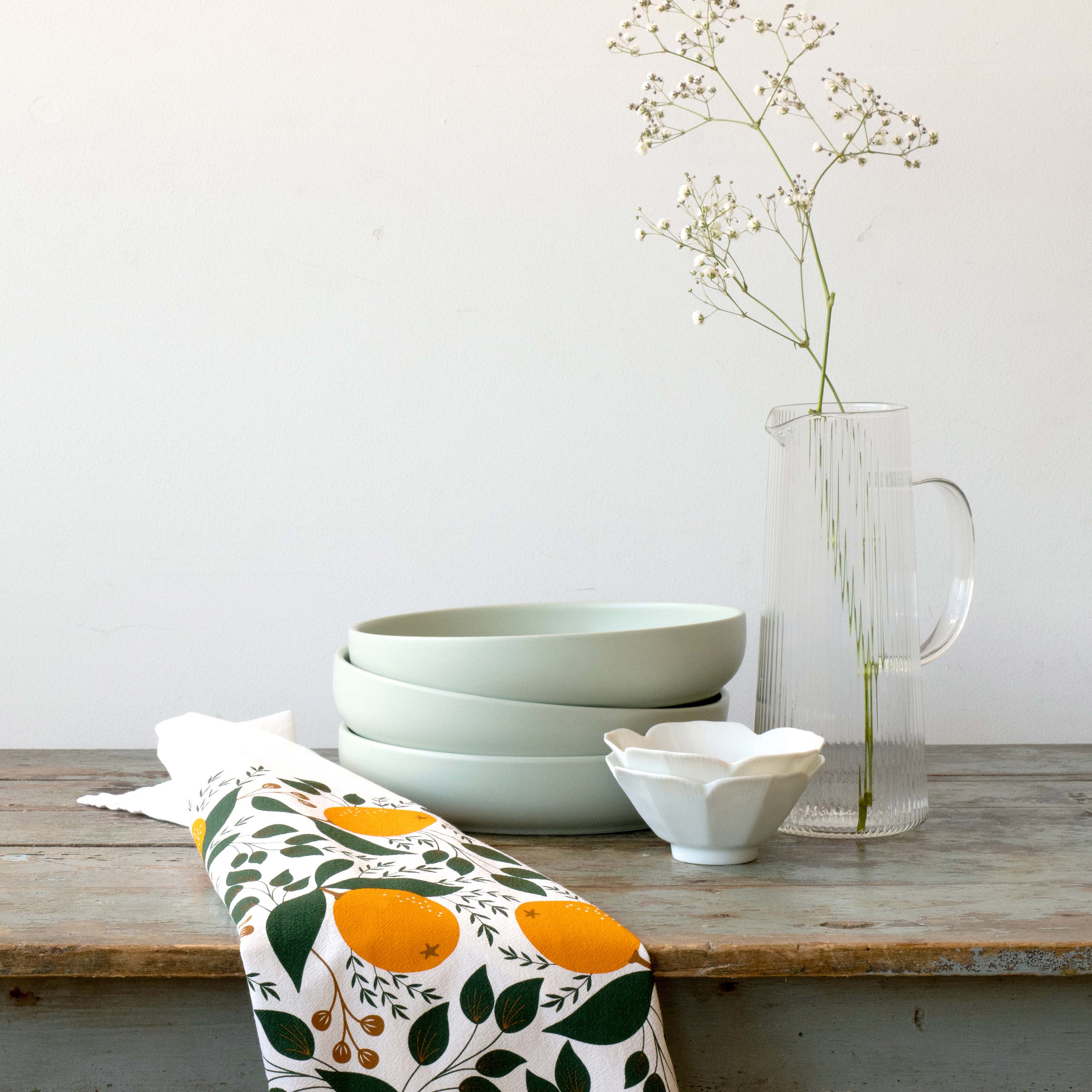 Winter Citrus Tea Towel styled beside stacked bowls and a glass vase on a wooden table
