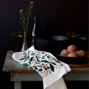 Winterberry Tea Towel styled on a rustic table with peaches, glassware, and dark cookware
