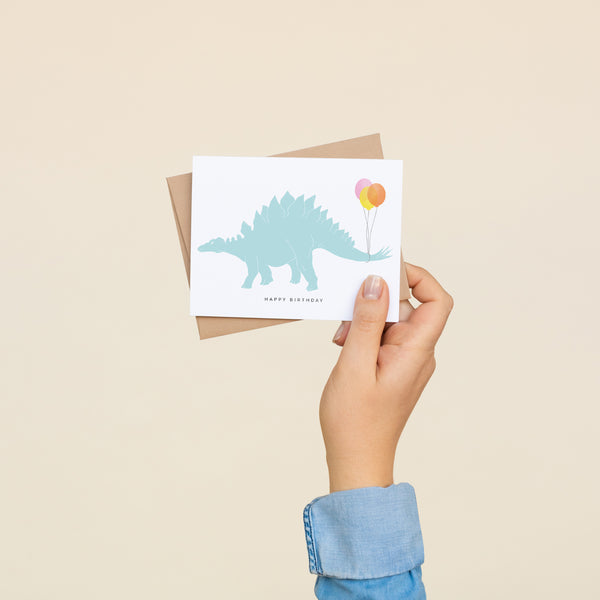 Single folded A2 greeting card with an envelope with an illustration of a blue dinosaur with three balloons wrapped around its' tail. Below the dinosaur is text that states "Happy Birthday".