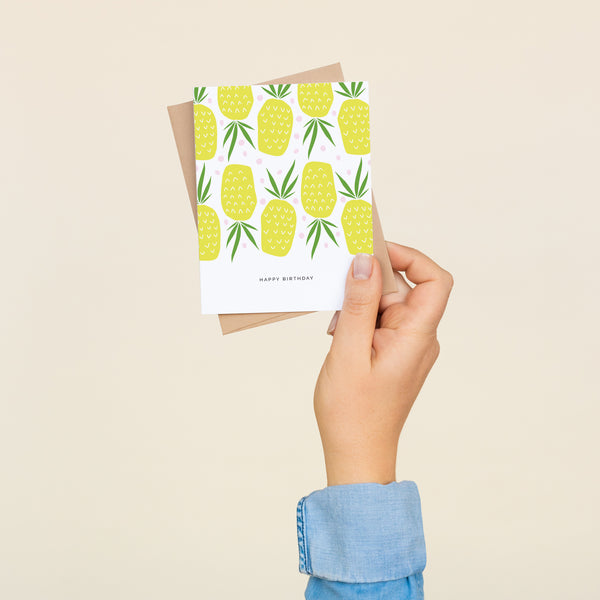 Single folded A2 greeting card with an envelope with an illustration of an alternating pattern of upright and upside-down pineapples. Below this illustration is text that states "Happy Birthday".