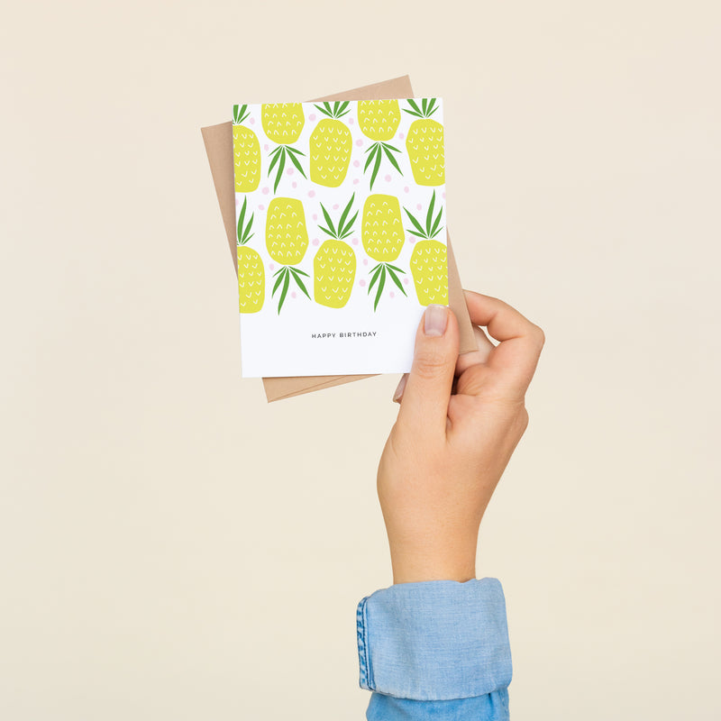 Single folded A2 greeting card with an envelope with an illustration of an alternating pattern of upright and upside-down pineapples. Below this illustration is text that states "Happy Birthday".