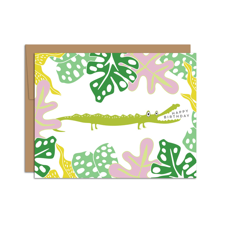 Single folded A2 greeting card with an envelope with an illustration of a crocodile in the center of the card surrounded by jungle leaves. The text "Happy Birthday" is coming out of the crocodile's mouth.