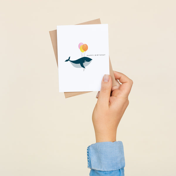 Single folded A2 greeting card with an envelope with an illustration of a whale holding three balloons. To the upper right of the whale is text that states "Happy Birthday."