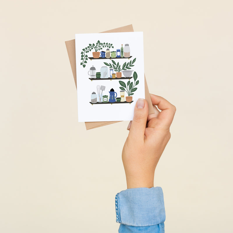 Box set of 8 folded A2 greeting cards with envelopes with an illustration of three shelves displaying various kitchen items such as a french press, mugs, and plants.