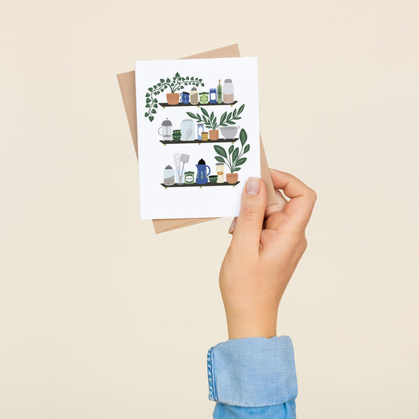 Single folded A2 greeting card with an envelope with an illustration of three shelves displaying various kitchen items such as a french press, mugs, and plants.
