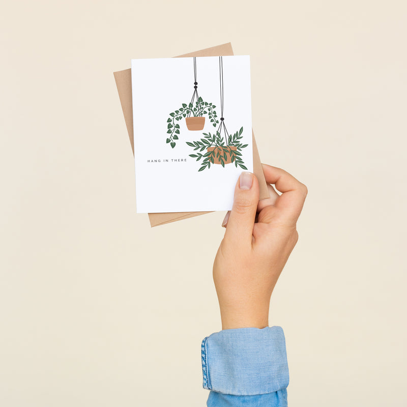 Single folded A2 greeting card with an envelope with an illustration of two potted plants hanging with text to the left that states "Hang in There".
