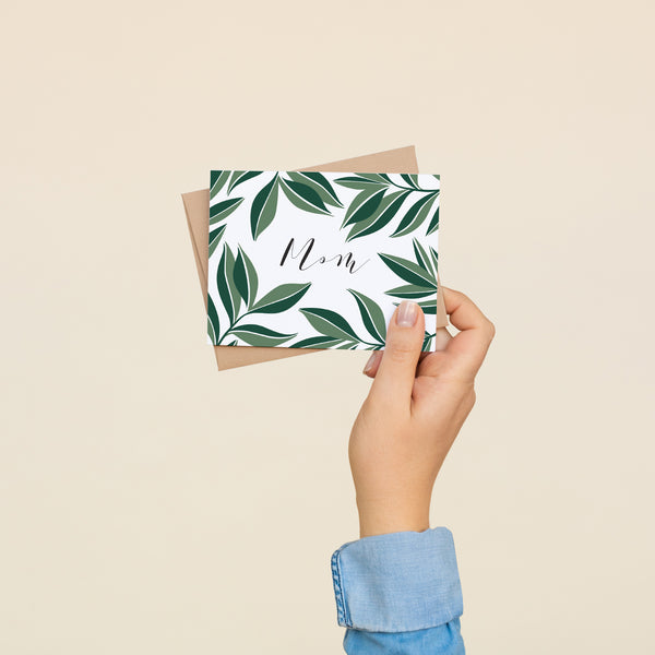 Single folded A2 greeting card with an envelope with an illustration of text stating "Mom" in cursive surrounded by dark green aster leaves.