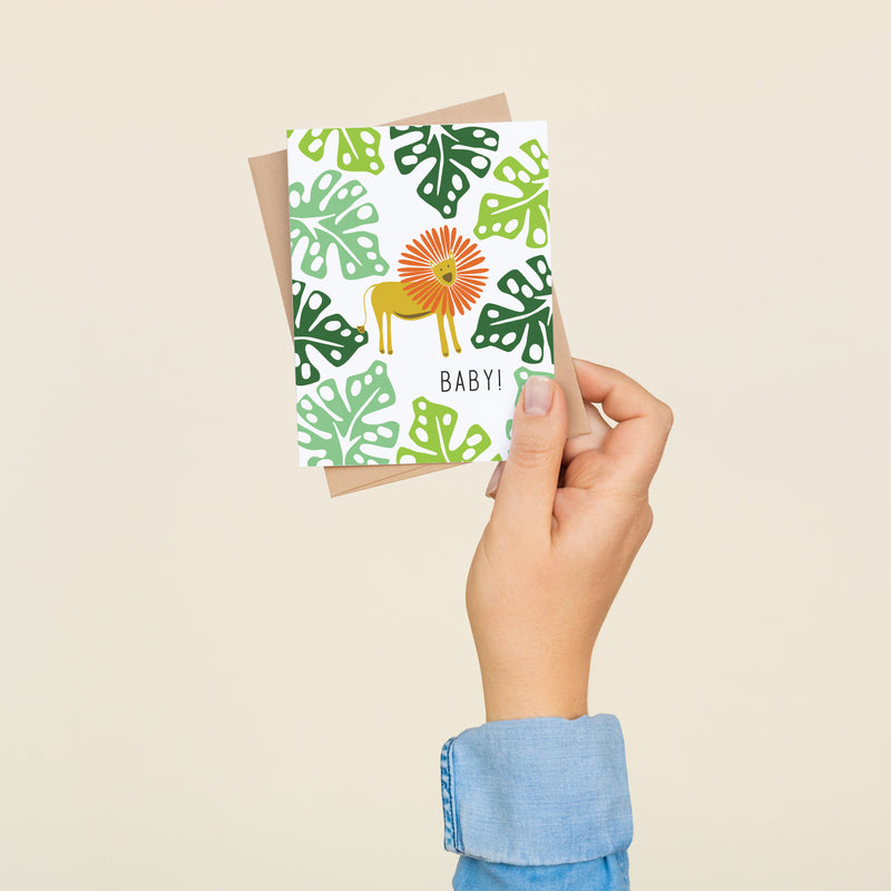 Single folded A2 greeting card with an envelope with an illustration of a lion surrounded by light and dark green jungle leaves. Below the lion is text that states "Baby!".