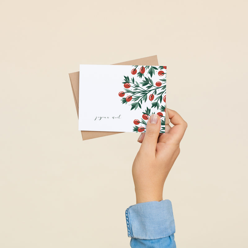 Box set of 8 folded A2 greeting cards with envelopes with an illustration  of red holly berries and green leaves bordering the right side of the card. On the bottom left side of the card is text in cursive that states "Joyeux Noel".