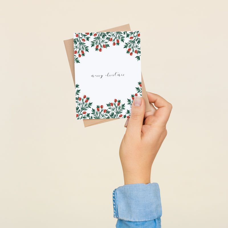 Single folded A2 greeting card with an envelope with an illustration of red holly berries and green leaves bordering the top and bottom of the card. In the center of the card is text in cursive that states "Merry Christmas".