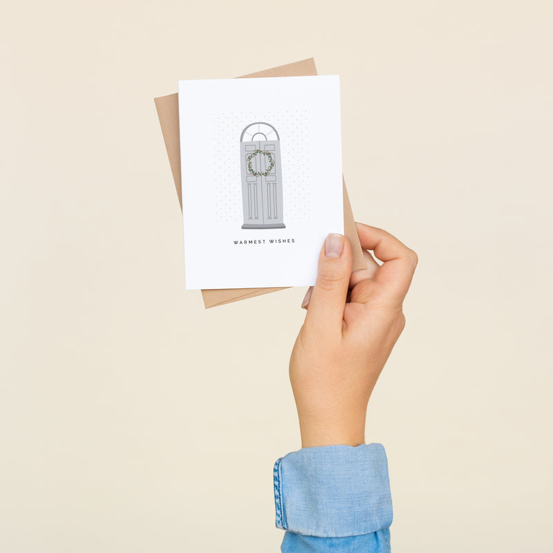 Single folded A2 greeting card with an envelope with an illustration of a gray door with a wreath and directly below it is text that states "Warmest Wishes".