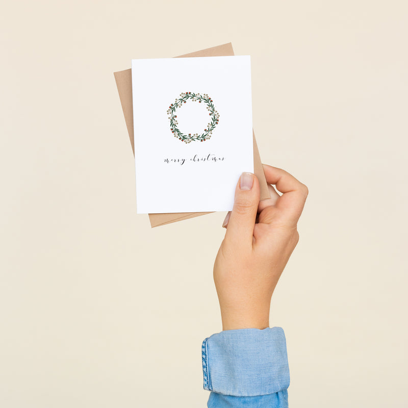 Box set of 8 folded A2 greeting cards with envelopes with an illustration  of a wreath centered in the card and text directly below it that states "Merry Christmas" in cursive.