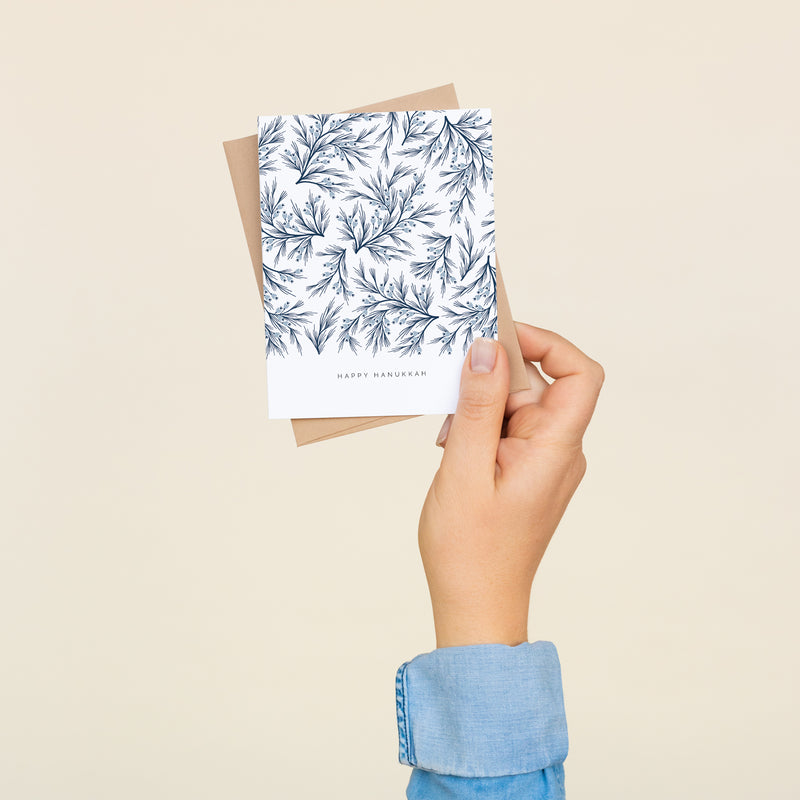 Single folded A2 greeting card with an envelope with an illustration of dark blue branches and light blue detail. Below this pattern of branches is text that states "Happy Hanukkah".