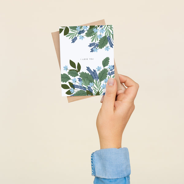 Single folded A2 greeting card with an envelope with an illustration of green leaves and blue flowers wrapping the top and bottom of the card. In the center is blank space with text that states "I Love You".