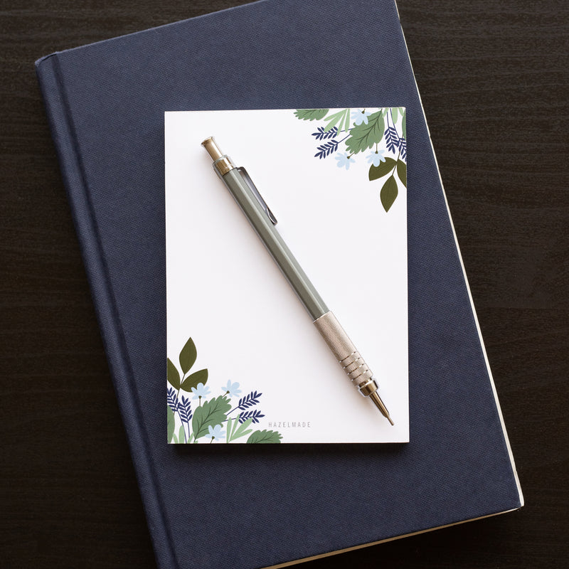 A single 4” by 5.5” small notepad with 50 tear-off sheets and an illustration of blue flowers and green leaves wrapping the top right and bottom left corner while the rest of the notepad is blank.