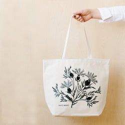 A single 18” by 15” by 7” natural cotton canvas tote bag with handle and an illustration of a branch of field thistle.