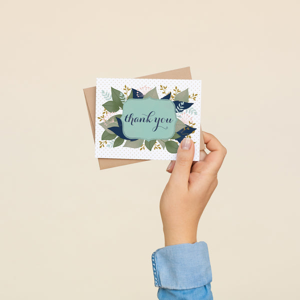 Single folded A2 greeting card with an envelope with an illustration of text stating "Thank You" in cursive bordered by a blue background and leaves sprouting behind it.