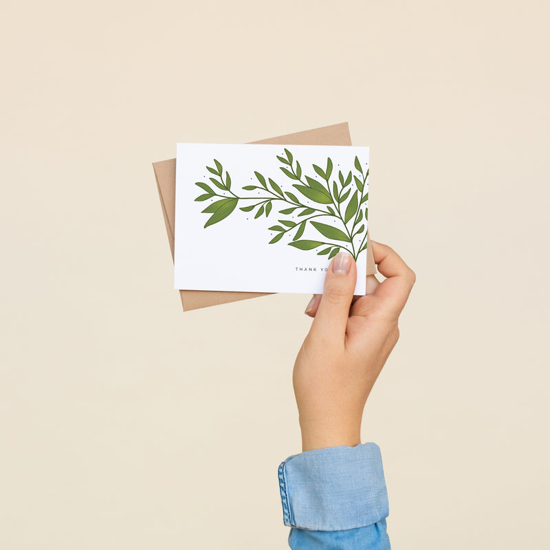 Single folded A2 greeting card with an envelope with an illustration of a green branch on the right side of the card and sprouting out towards the left. Below the branch is text that states "Thank You".