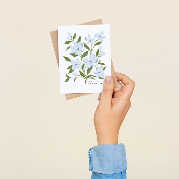Box set of 8 folded A2 greeting cards with envelopes with an illustration   of blue dogwood flowers and green leaves. Text below and to the right of the bouquet states "Thank You" in cursive.