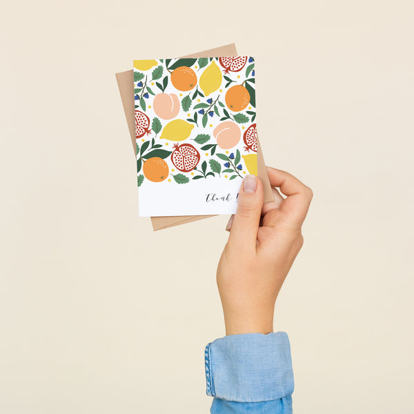 Single folded A2 greeting card with an envelope with an illustration of pomegranates, lemons, oranges, blueberries, and peaches in a mixed pattern. In the bottom right is text in cursive that states "Thank You".