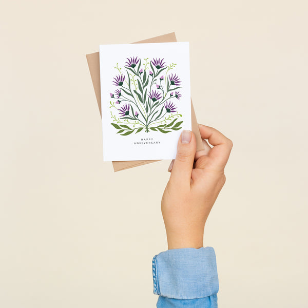 Single folded A2 greeting card with an envelope with an illustration of purple thistle and green leaves with text below it that states "Happy Anniversary".