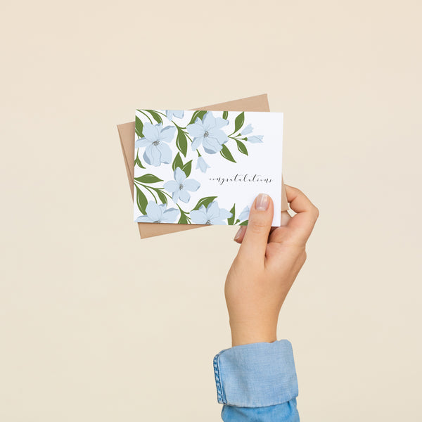 Single folded A2 greeting card with an envelope with an illustration of blue dogwood flowers wrapping the top, bottom, and left side of the card. On the right side is text that states "Congratulations".