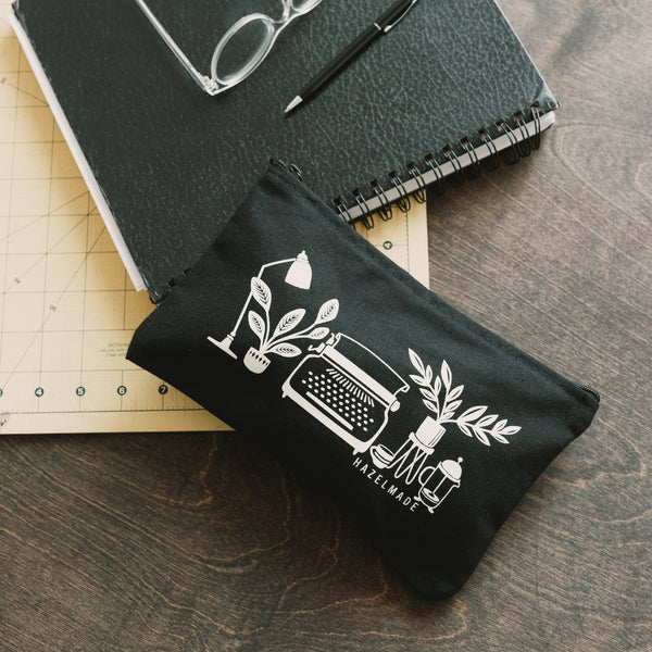 A single 9” by 6” black cotton flat zip pouch with an illustration of a typewriter and other desk items.